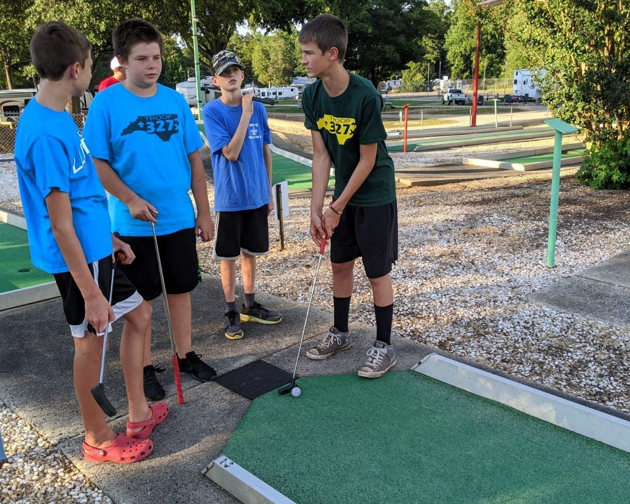 Four boys in athletic shirts stand on a mini golf course, talking beside a putting green.