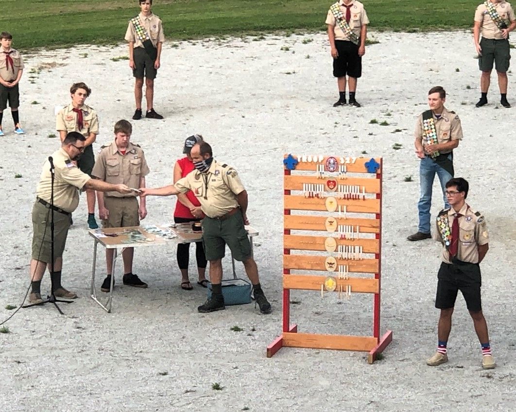 People in beige uniforms around a table outdoors, with a colorful board and several onlookers nearby.