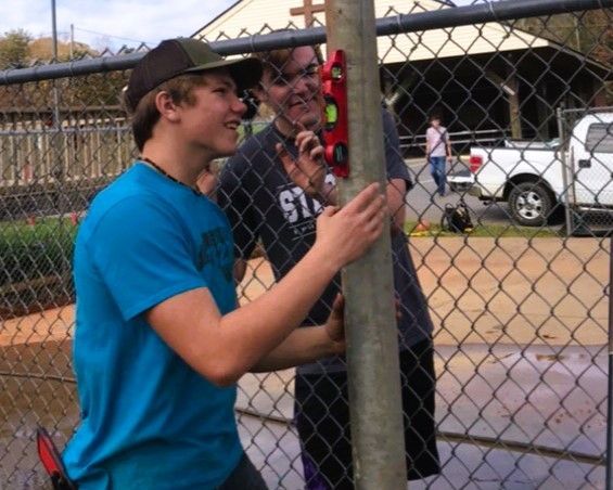Two people smiling by a chain-link fence at an outdoor event, one holding a wooden post.