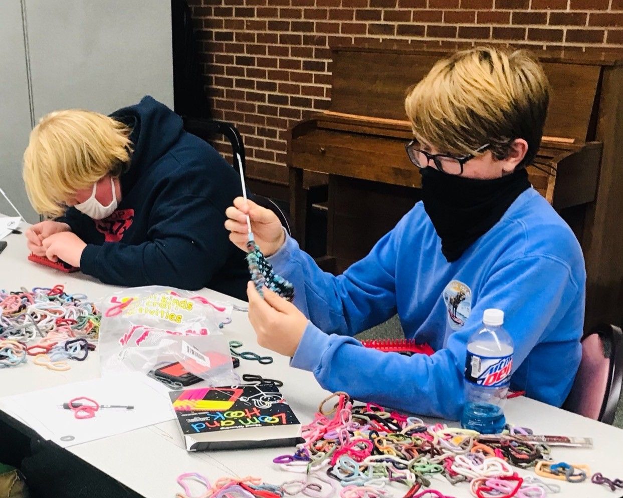 Two masked people making beaded crafts at a table in a brick-walled room