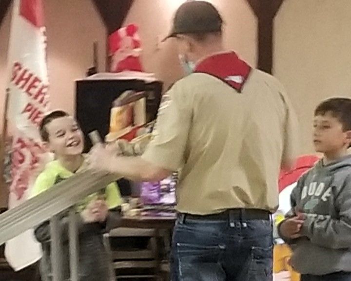 Boy handing an item to a scout leader while another child watches indoors