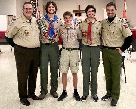 Five scouts in tan and green uniforms standing indoors, smiling with hands around each other.