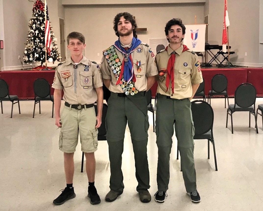 Three scouts standing in a hall, wearing tan and green uniforms with neckerchiefs and badges.