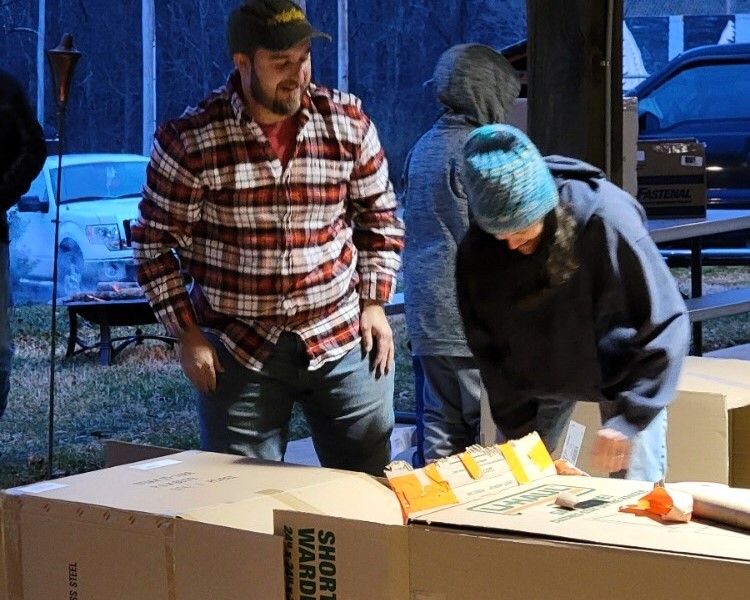 Two people packing boxes at a table outdoors, one in a plaid shirt and one in a blue beanie.