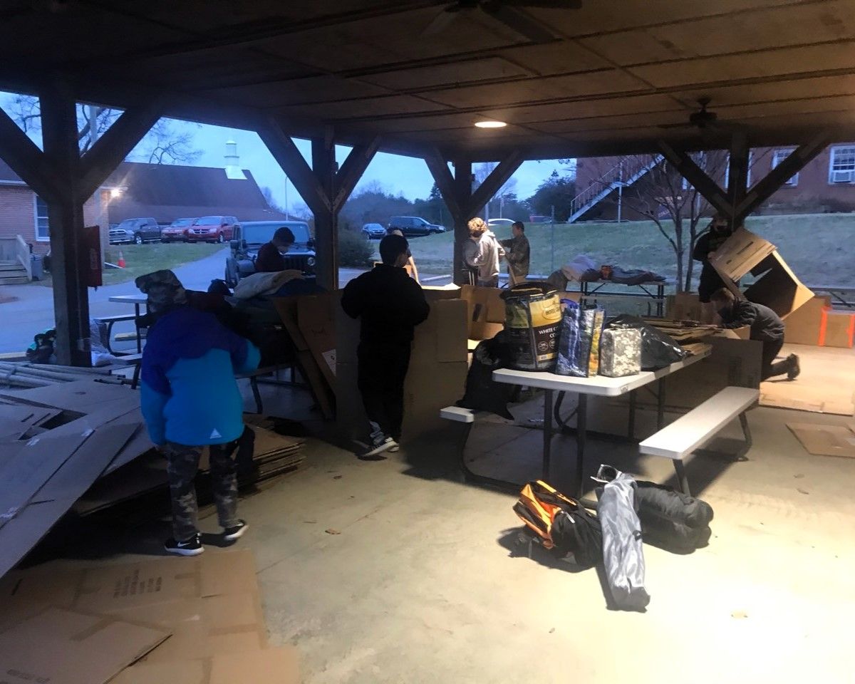 People gathered under a pavilion around a grill and tables at dusk, with coats on and bags nearby.