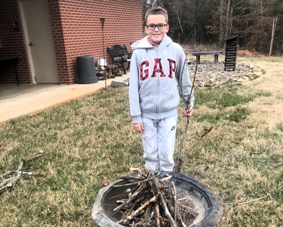 Person stands outdoors near a fire pit with sticks, wearing a gray GAP hoodie and sweatpants.
