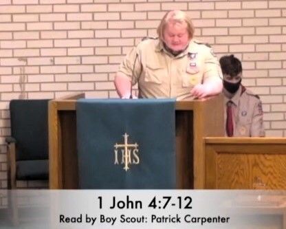 Boy Scout reading at church podium beside a man, with caption “1 John 4:7-12”