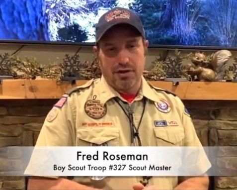 Boy Scout Troop #327 Scout Master Fred Roseman in tan uniform and cap, standing indoors before a nature backdrop