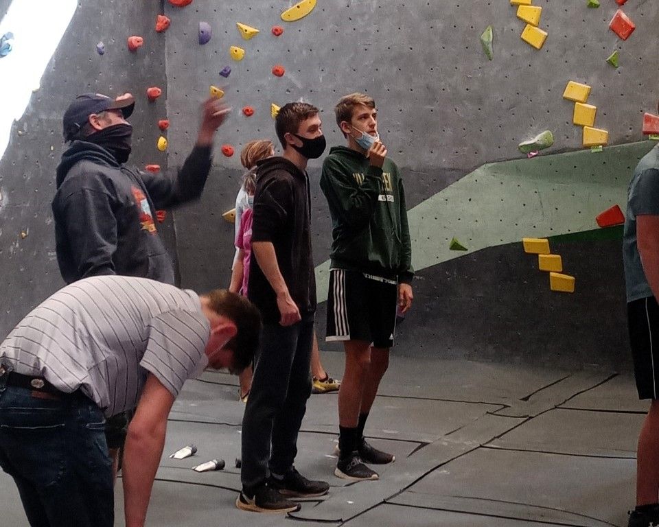 People bouldering indoors on a climbing wall, with colorful holds and several climbers waiting nearby.