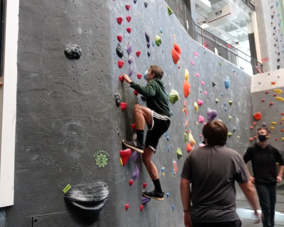 People rock climbing indoors on a gray wall with colorful holds and belayers below