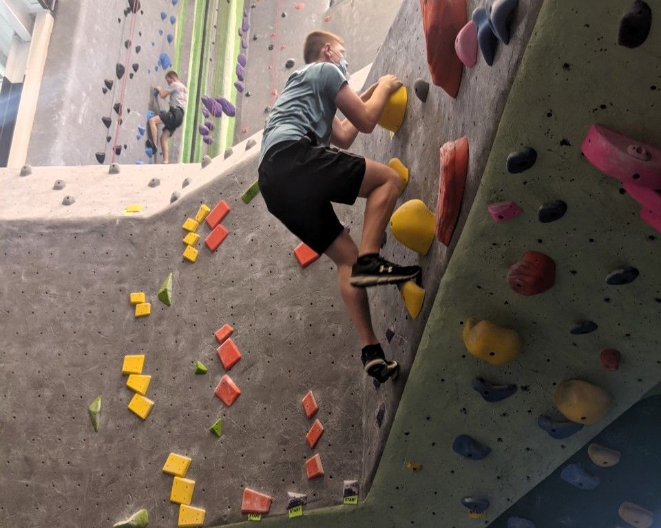 Person rock climbing indoors on a colorful climbing wall with yellow, pink, and orange holds.