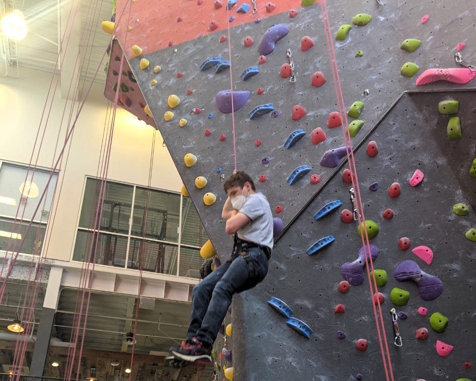 Person climbing an indoor rock wall with colorful handholds, wearing a harness and helmet