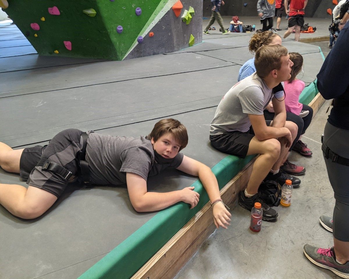 People resting at an indoor climbing gym beside a gray wall with green holds.