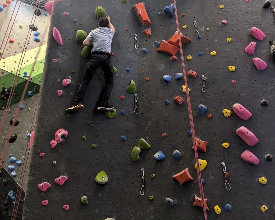 Person climbing an indoor rock wall with colorful holds and safety ropes.