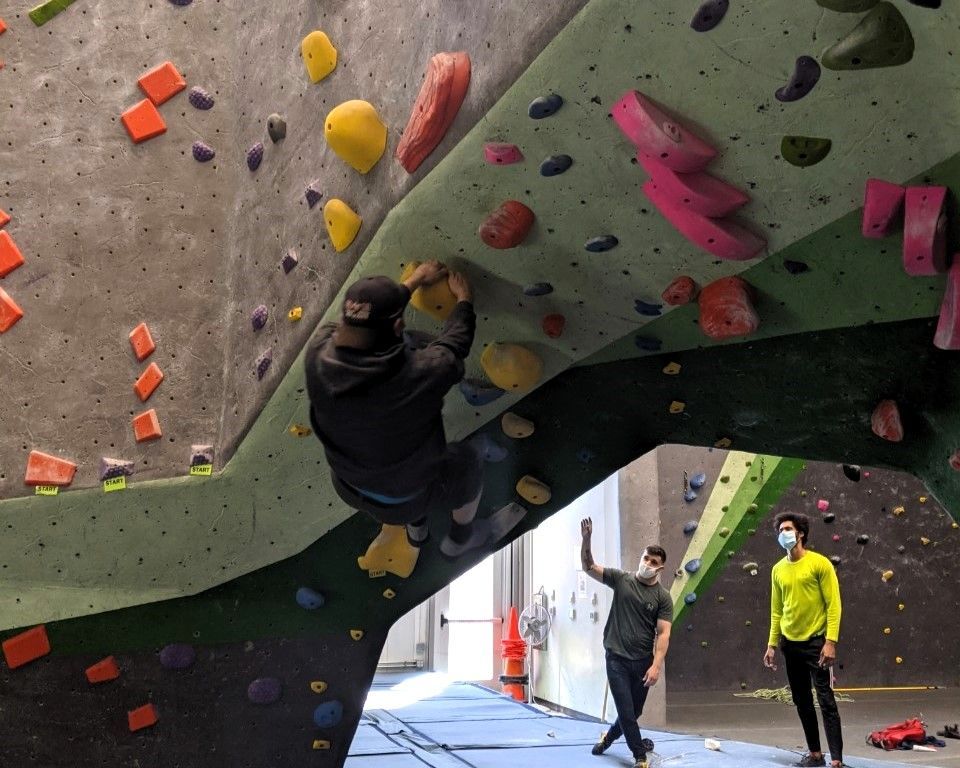 People bouldering indoors on a colorful climbing wall, with one climber hanging under an overhang.