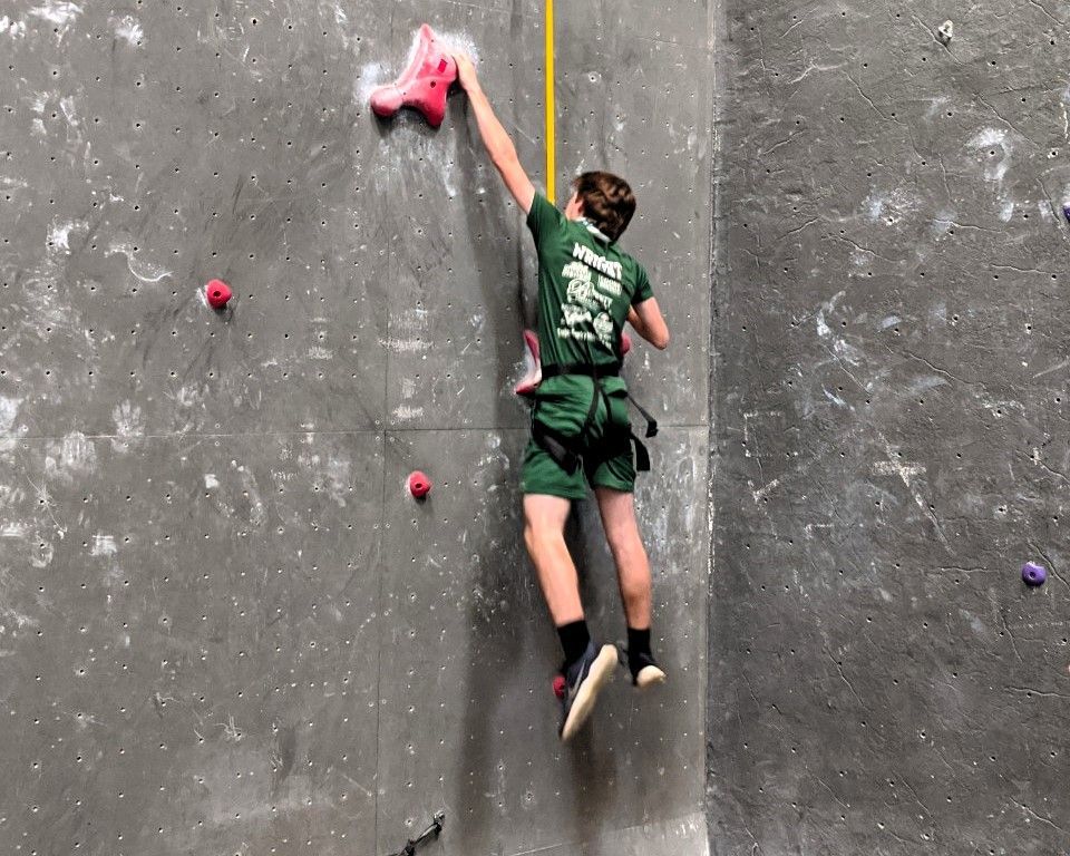 Rock climber in green reaches for a pink hold on a gray indoor climbing wall