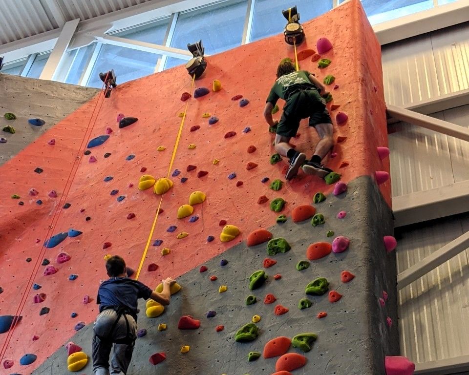 Indoor rock-climbing wall with several climbers on an orange wall with colorful holds.