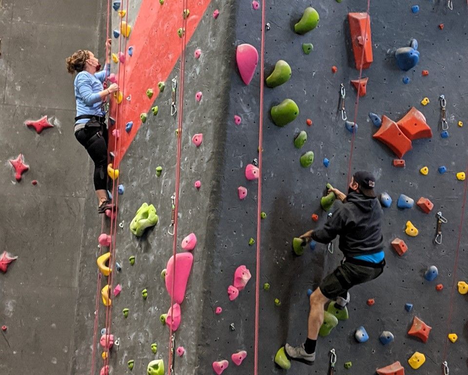 Two climbers on an indoor rock wall with colorful holds, one in pink and one in black.