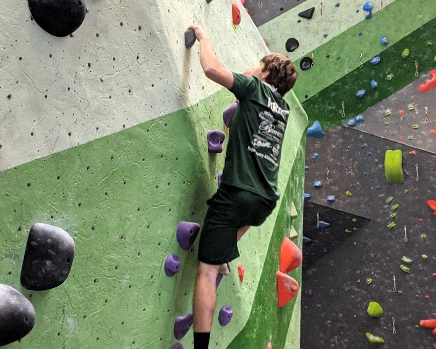 Indoor rock climber on a green-and-gray wall, reaching for a hold with colorful holds nearby