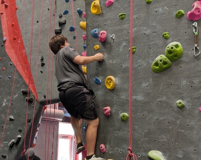 Person climbing an indoor rock wall with colorful holds and ropes attached