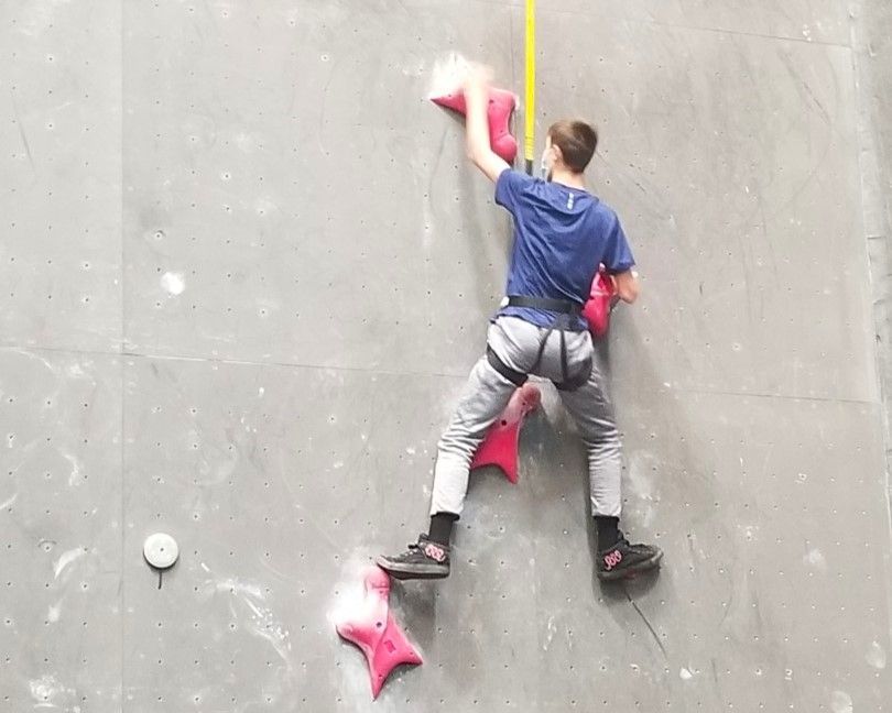 Person rock climbing on a gray wall with pink holds, reaching upward.
