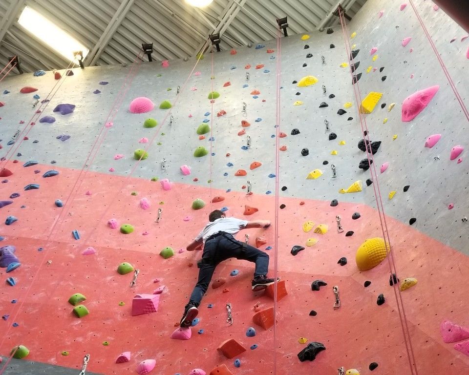 Person climbing an indoor rock wall with colorful holds and ropes