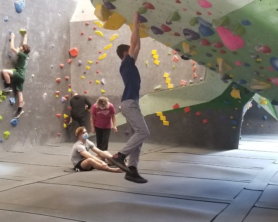 Indoor bouldering gym with climbers on a colorful wall and padded floor; one person reaching overhead.
