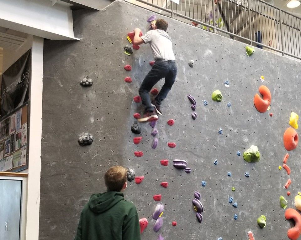 Indoor climbing wall with a person climbing red holds while another watches below.