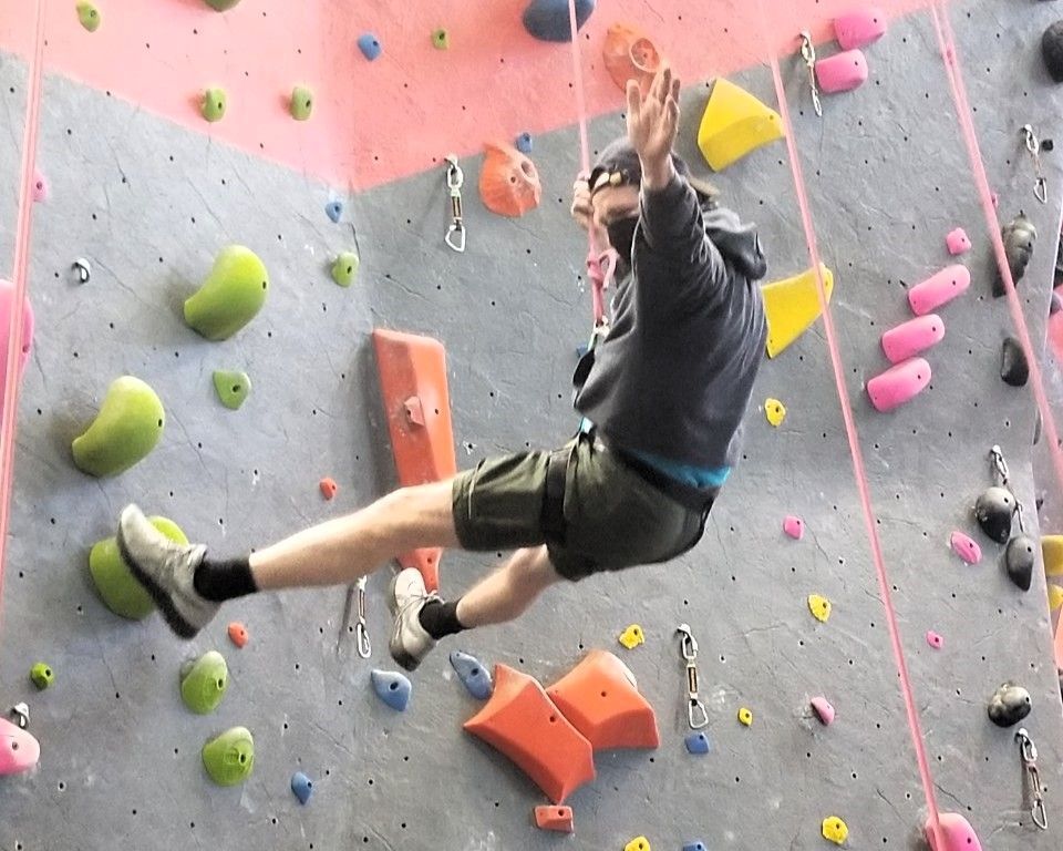 Person rock climbing on an indoor wall with colorful holds, one leg extended and clipped to a rope.