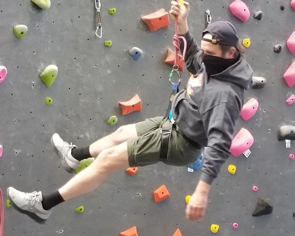 Climber on indoor bouldering wall, hanging from a rope against gray wall with colorful holds.