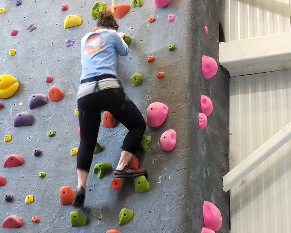 Person rock climbing on an indoor wall with colorful holds, reaching upward.