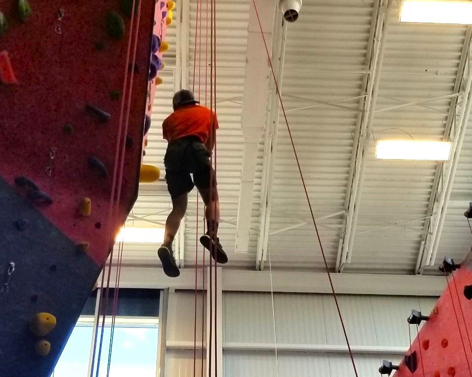 Person rock climbing indoors on a red climbing wall, wearing an orange shirt and black shorts.