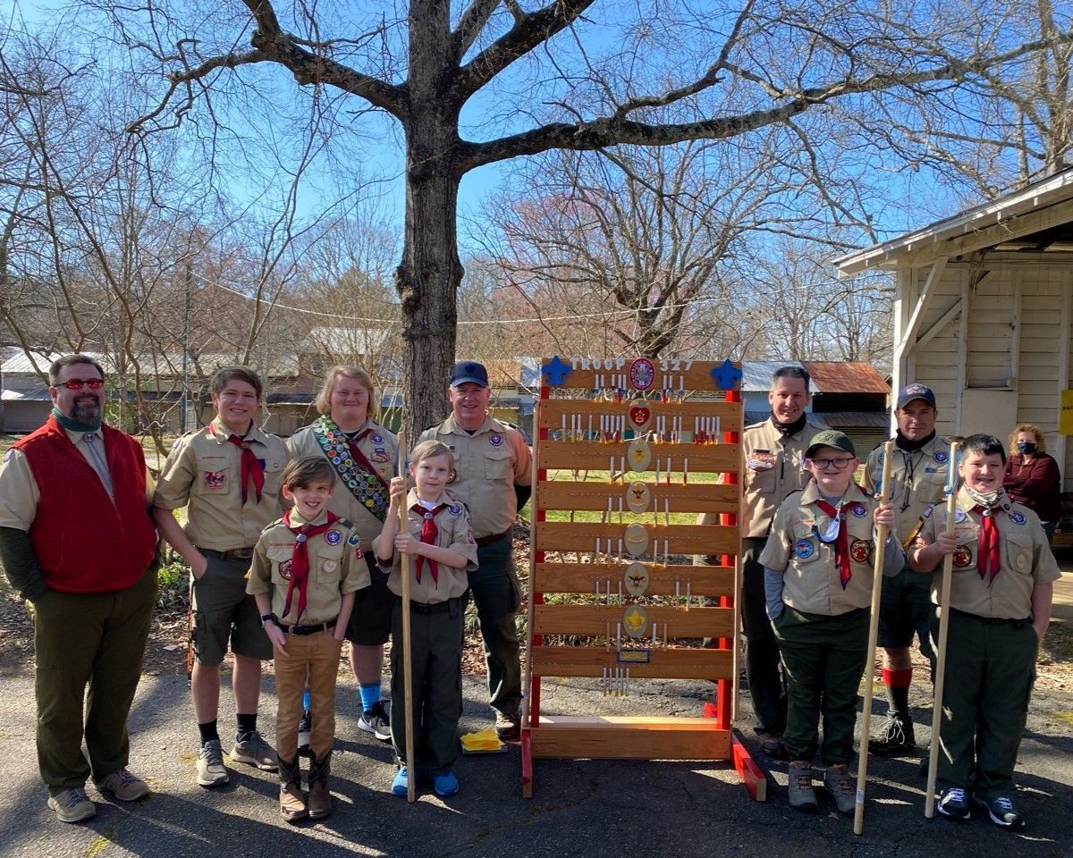 Group of scouts and leaders posing outdoors beside a wooden award display on a sunny day