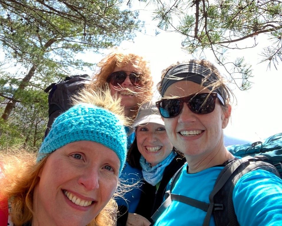 Four smiling hikers in bright outdoor gear taking a selfie among pine trees