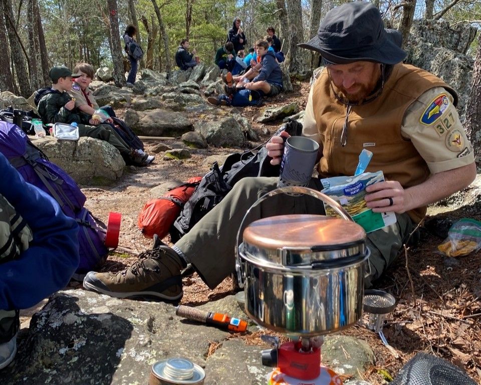 Hikers resting in the woods, sharing food beside a camp stove and cookware.