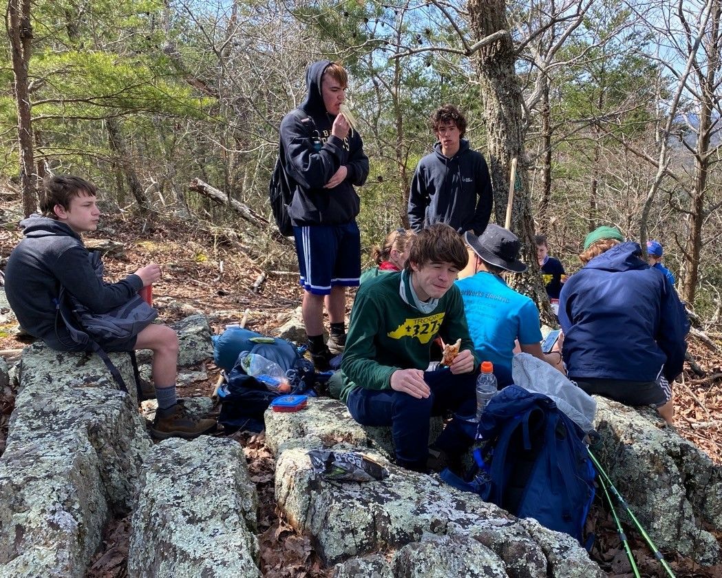 Group of hikers resting on rocky outcrop in a wooded area, with backpacks and outdoor gear.