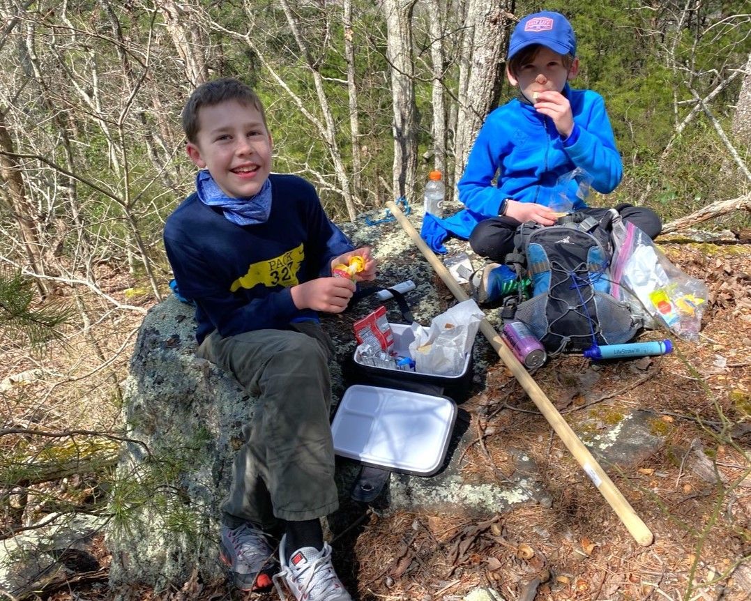 Two children sit on rocks outdoors with a pile of trash bags and a rake, cleaning up a wooded area.