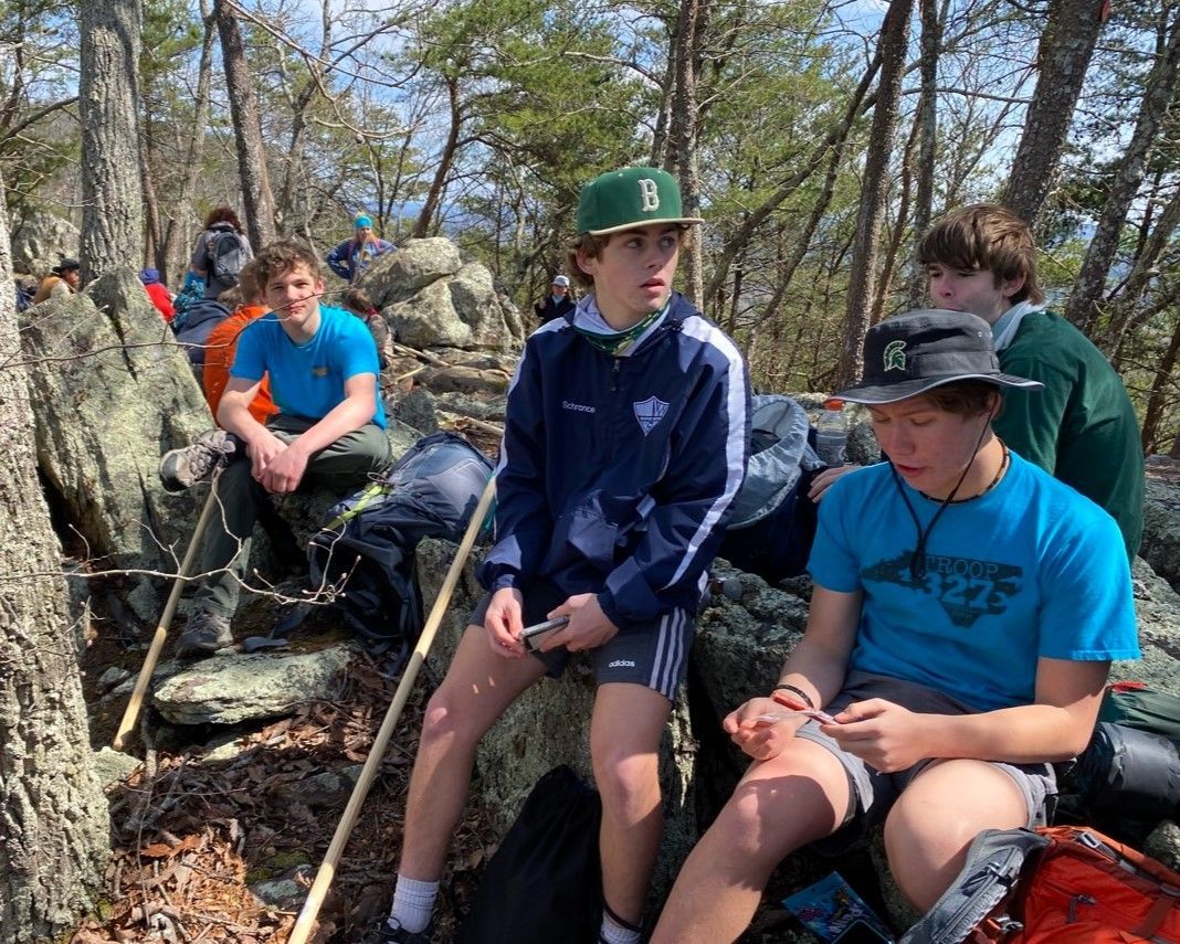 Group of hikers resting on rocky trail in a wooded area, with two sitting in the foreground.
