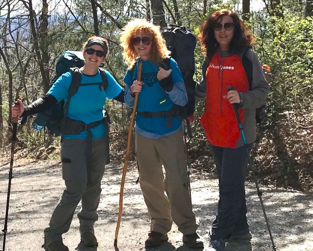 Three hikers with backpacks and walking poles on a wooded trail, smiling in the sun