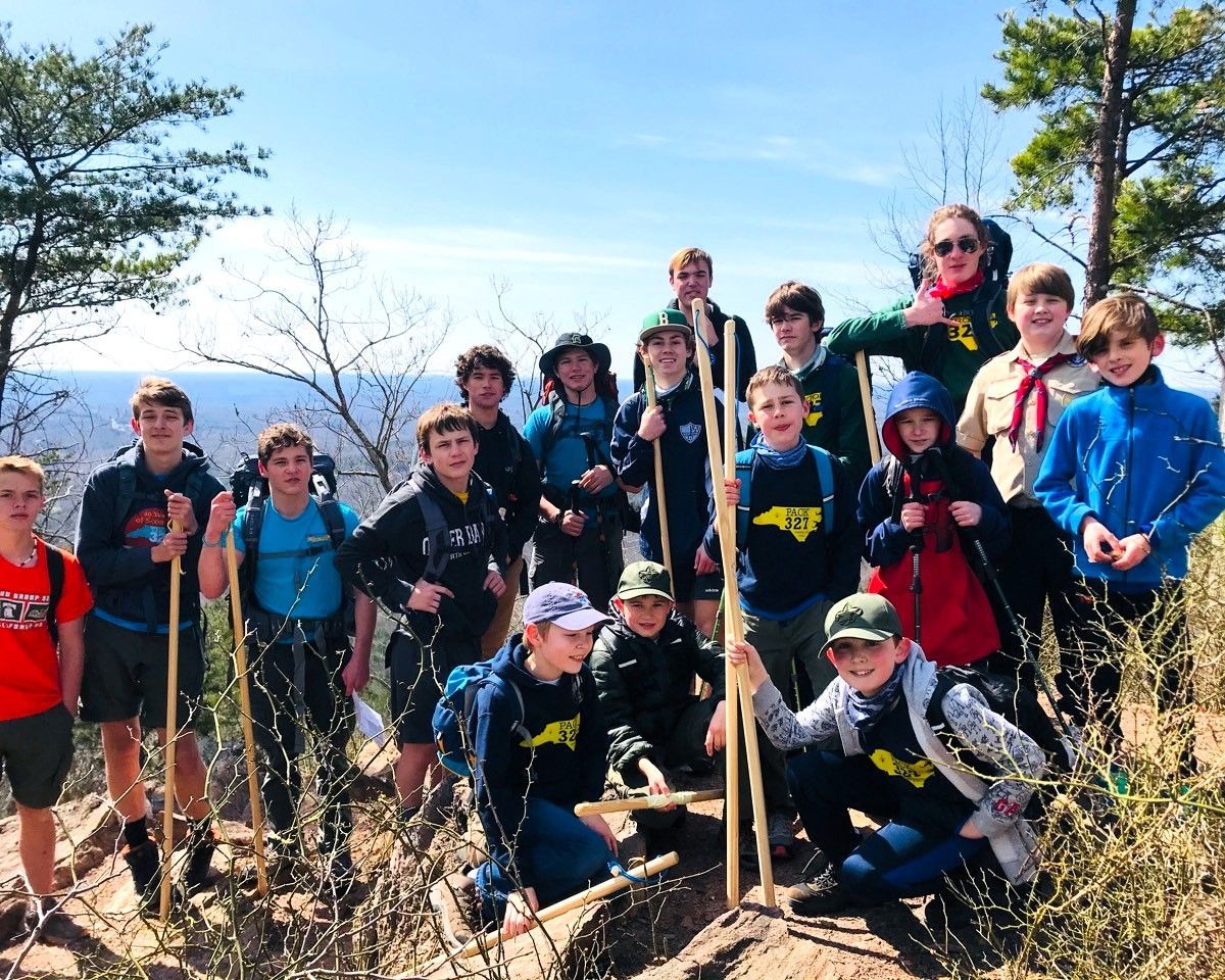 Group of hikers posing on a rocky mountain overlook with trees and distant hills under a blue sky