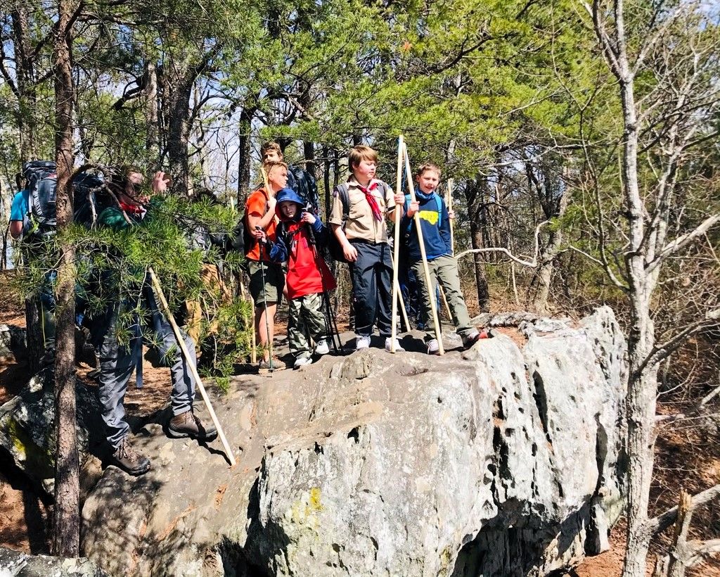 Hikers standing on a rocky cliff in a wooded area, wearing backpacks and holding trekking poles.