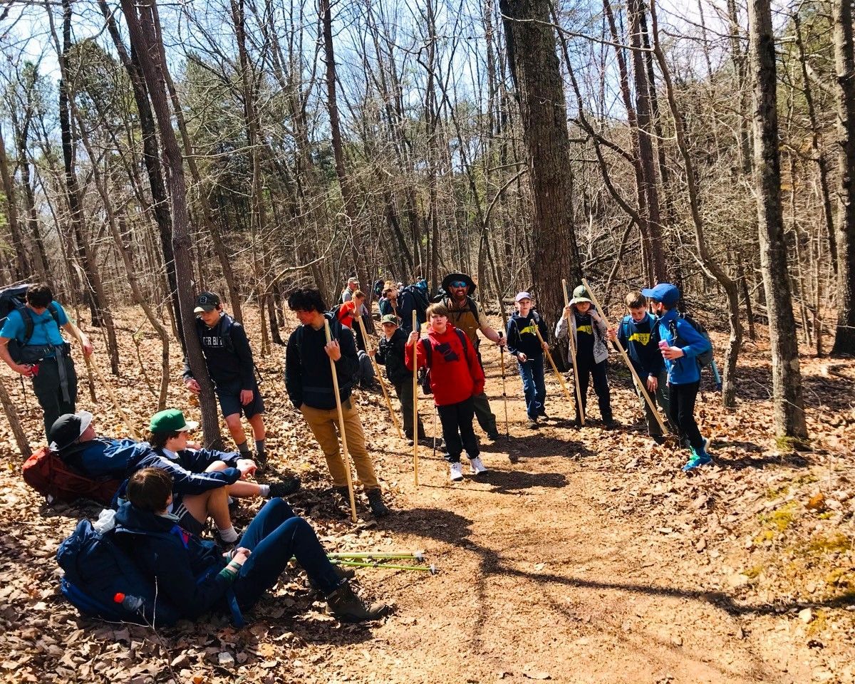 Group of hikers with backpacks on a wooded trail, some standing and some sitting in sunlight.