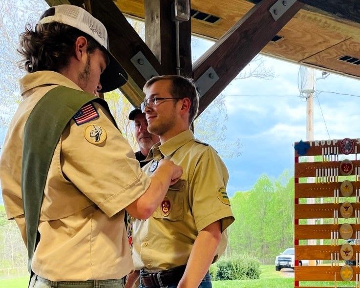 Two Scouts in tan uniforms speak under a wooden pavilion, with a rack of badges and blue sky behind them.