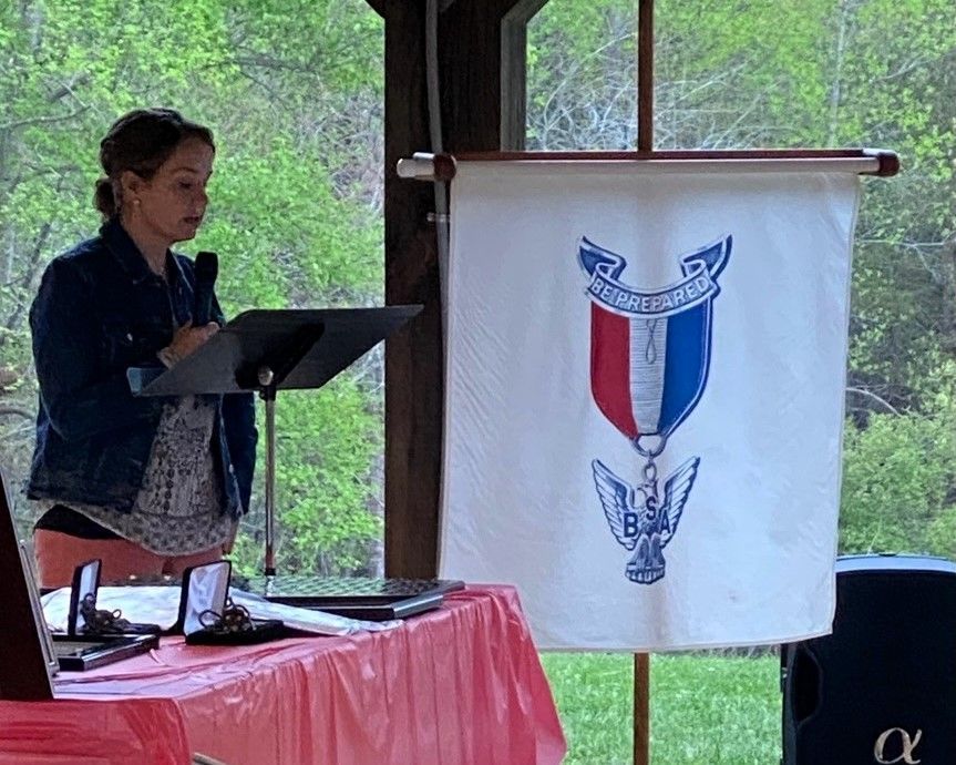 Woman speaking at a podium beside a red-white-blue banner in a covered outdoor pavilion