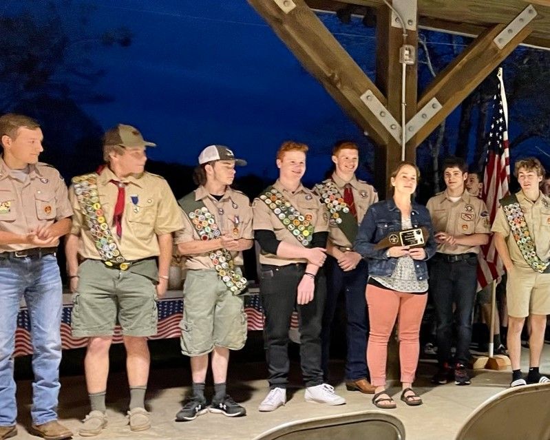 Scout troop standing on stage holding trophies under wooden beams and an American flag at night