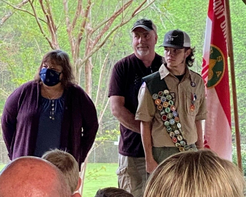 Three people standing outdoors under a pavilion, with a scouting sash and red flag visible.