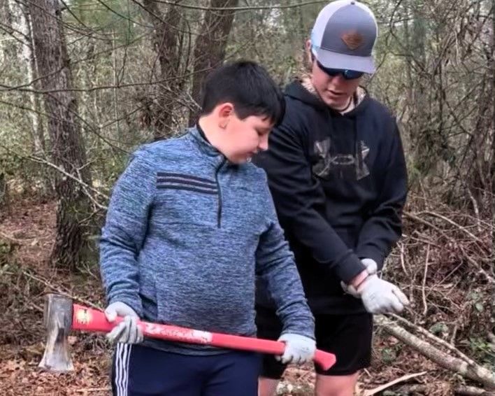Two people in a wooded area holding an axe and gloves, appearing to clear branches together