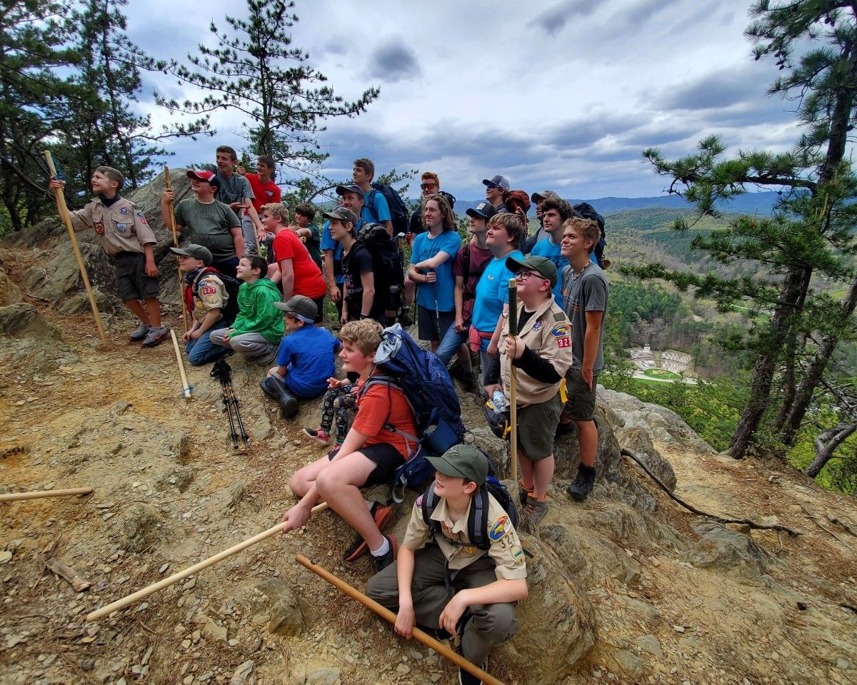 Group of hikers posing on a rocky mountain trail under cloudy skies, surrounded by trees.
