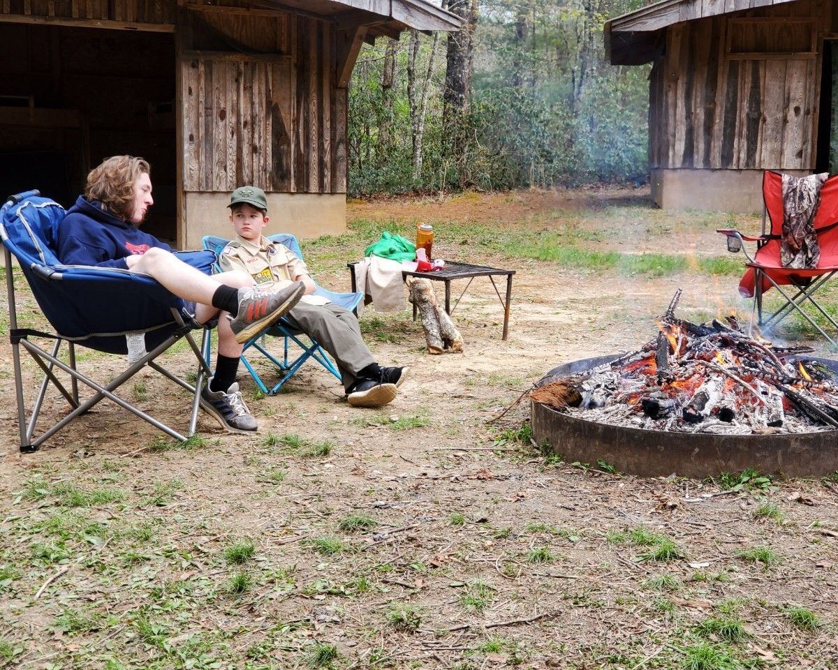 People relaxing in camp chairs by a fire pit at a rustic outdoor campsite.