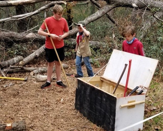 Three people in a wooded area using tools around an open chest or bin filled with items.